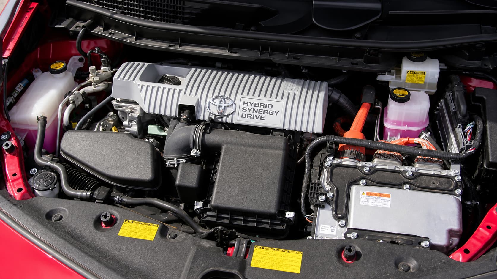 Compartiment moteur ouvert de la Toyota Prius 5, dévoilant le couvercle en plastique marqué Toyota Hybrid Synergy Drive, cerné de câblages et conduits ordonnés.